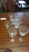 Photo of nine glasses including a small brandy glass and eight taller floral etched crystal glasses arranged on a wood table.