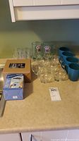 Lot contents on countertop including glass steins with A&W logos, coffee mugs, bowls, small glasses, and boxed kitchen scale.