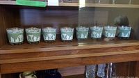 Eight Grecian-style glasses arranged in a row on a wooden shelf with a glass front. Each glass features white figure designs on a green background with gold trim around the top.