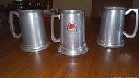 Three pewter mugs on a wooden surface, two plain and one with a red and white label on the front.