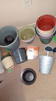 Collection of various plant pots of different colors and materials arranged on floor, metal, plastic, and ceramic visible