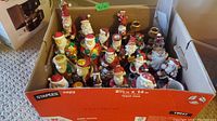 Box containing multiple Santa Claus figurines, candle holders, and helpers. Different sizes and colors mostly red, white, green, and gold.