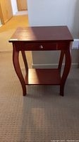 Front view of wooden side table showing drawer with round knob and lower shelf, standing on carpeted floor