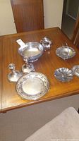 Photo of the silver plate set on a wooden table showing multiple pieces including large bowl, pitcher, small dishes, and candle holders
