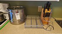 Photo showing stainless steel stock pot, metal wire organizer rack, and wooden chef knife block with knives on kitchen counter.