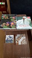 Shows two hardcover books: Anne Geddes 'A Labor of Love' featuring babies with floral hats, and Robert Bateman 'Birds' with a white crane on the cover, along with two small square decorative trivets placed on a wooden table.