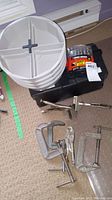 Photo showing black toolbox, drill bits set, several white compartmentalized bucket trays, 3 metal C-clamps and one pair of locking pliers on carpet.