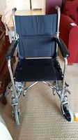 Front view of black transport wheelchair with nylon seat, padded armrests, and chrome frame on carpeted floor.
