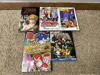Six anime and video game themed manga and comic books arranged on carpet: Zelda Twilight Princess, Boruto, Dragonball Super, Sonic Select, Pokémon Black & White Vol. 1