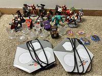 Photo showing a Disney Infinity figure lot including various detailed character figures such as Iron Man, Sulley, Captain America, Lightning McQueen, Mater, Incredibles characters, and Jack Skellington. Two Disney Infinity base plates and several clear bases and game discs are visible in front.