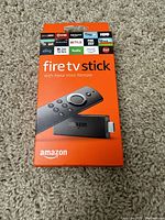 Front of Amazon Fire TV Stick box displaying device image, Alexa Voice Remote and streaming service logos.