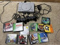 Full lot arranged on floor including PlayStation 1 console, two controllers, and nine game discs in cases plus two loose discs.