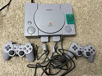 Front view of PlayStation 1 console with two controllers and green memory card attached, showing power and reset buttons.