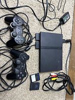 Full view of PlayStation 2 Slim console with two wired DualShock controllers, two memory cards, and AV cables on carpet.