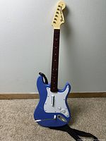 Front view of blue Fender wireless guitar controller for PS3 Rock Band with strap attached, showing the white pickguard and control buttons.