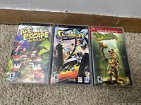 Front view of three PSP game cases: Ape Escape On the Loose, GripShift, and Daxter Greatest Hits placed on carpeted floor against a wall.
