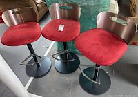 Three metal and upholstered barstools with brushed metal backrests and red cushioned seats arranged together on a concrete floor.