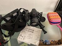 Camera with lens mounted, black camera bag on left, user's manual beneath camera, small colorful underwater camera case on right.