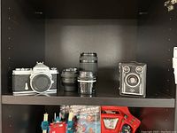 Overall shelf view showing Nikon Nikkormat body at left, two lenses at center, Agfa Synchro Box camera at right