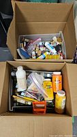 Two open cardboard boxes filled with assorted paint bottles, brushes, and craft materials seen from above.