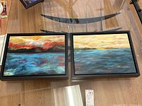 Pair of framed artwork prints on a wooden table showing seascape scenes with colorful skies, black frames, and visible artist signature.