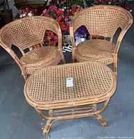 Set of one oval rattan coffee table and two rounded back rattan chairs with woven rattan seats and backs, displayed together on dark floor.