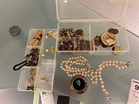 Overview of plastic compartment box holding multiple small religious items, rosaries, crosses, medals and coins laid out on glass surface