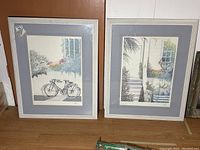Photo of two framed artworks showing a bicycle and a porch scene by Heine, sized 24 x 31 inches.
