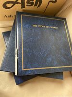 Two volumes of "The Story of Canada" stamp collections showing dark blue binders, one with gold embossed title.