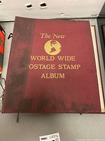 Front cover of the stamp album showing the title 'The New World Wide Postage Stamp Album' embossed in gold on a maroon hardcover.
