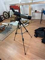 Spotting telescope mounted on adjustable tripod on wood floor, with art and other items in background.