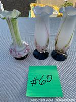 Photo showing three tall glass vases with ruffled rims, white with green and pink accents, standing on a gray surface outdoors.