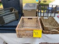 Full view of various woven baskets and bins including a large fabric-lined wicker basket, different wicker tray and basket styles, and black woven trays stacked.