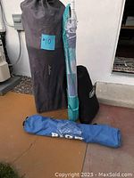 Four camping and beach items standing upright in bags on a hard surface: a tall grey sleeping bag, turquoise and white beach umbrella, black storage bag likely for chair, and blue bag for single chair.