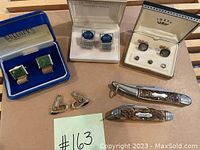 Overview of all items — cuff links in boxes and two folding pocket knives on cardboard