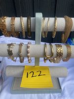 Multiple gold toned bracelets displayed on a light gray bracelet stand, showing variety of styles and textures.