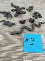 Nine assorted resin decor figurines including lizards, frogs, snake, and toad on beige woven mat.