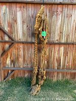 Two long handmade macrame plant hangers hanging against a wooden fence outdoors. The hangers are made from natural beige rope with decorative knots and tassels. Green tag labeled '112' attached to one hanger.
