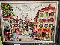 Framed needle point artwork showing a colorful town street scene with several multi-story buildings including a boulangerie and café, people walking, greenery, and a domed building in the background.