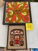 Two framed vintage needle point decor pieces displayed, one larger sun design and one smaller abstract face design.