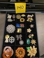 Full view of all vintage pins and brooches arranged in a black divided tray showing various designs and colors.