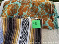 Two folded textiles including a large floral patterned bedspread in rust and blue, and a striped Mexican serape style blanket with fringe.