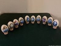 Set of 10 porcelain eggs with sea artwork displayed in a row on black fabric background showing fronts and stands. One stand has chip on edge.