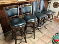 Four bar stools: two wooden stools with black cushioned seats and backs, two taller metal stools with ribbed black vinyl upholstery, arranged in a row alongside a bar counter.