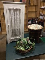 White wooden corner cabinet with lace curtain door, barrel-style wooden stool with floral cushioned seat, decorative basket with artificial plant, and dried plant, all displayed together.