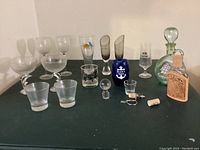 Photo of 18 assorted barware items including dolphin stem wineglasses, cocktail glasses, shot glasses, two decanters, corks, and accessories arranged on a dark surface.