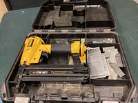 Top view of DeWalt yellow air nailer with black handle and trigger, placed inside open black plastic carrying case with multiple stacks of rectangular brad nail strips on the right side.