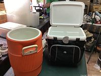 Two coolers side by side on a table. Orange barrel style cooler with white interior and handles. Black rectangular cooler with white lid and zippered soft pouch on front. Both lids open showing interior.