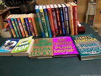 Photo showing all Janet Evanovich books arranged on a table, including paperback and hardback editions and extra books spread in front.