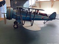 Side view of the vintage Japanese tin biplane showing the sky blue body, propeller, wheels, and pilot figure inside the cockpit. The model has metal frame support between the wings.